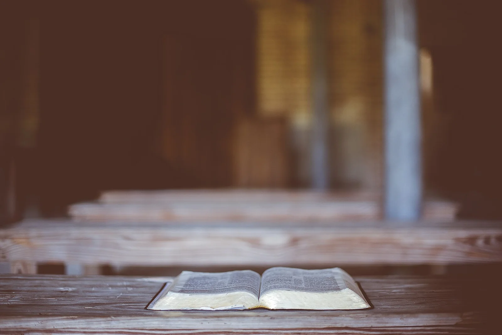 An open Bible resting on a wooden church pew, with empty pews softly out of focus behind it