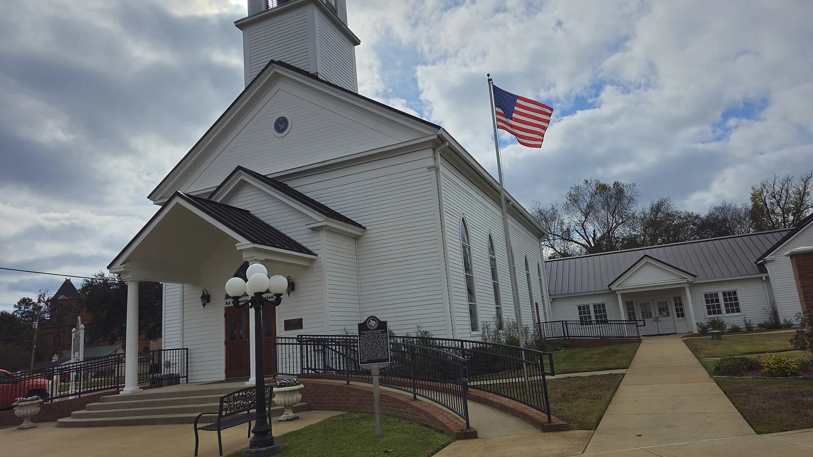 White clapboard Catholic church with a steeple, covered entrance and a railed wheelchair ramp leading to the door